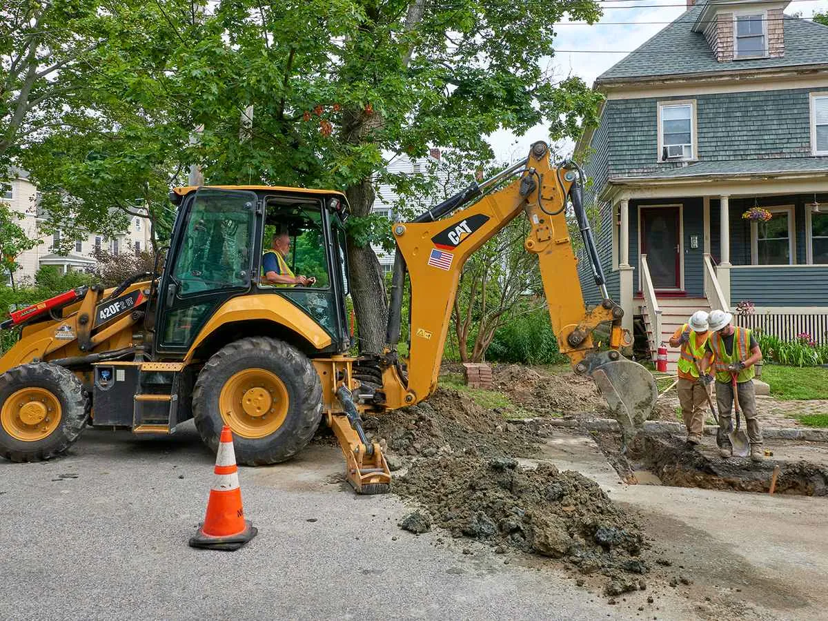 A yellow CAT backhoe loader is digging a utility trench on a residential street in front of a blue house. The operator is in the cab, while two construction workers in hard hats and safety vests stand near the trench, guiding the excavation.
