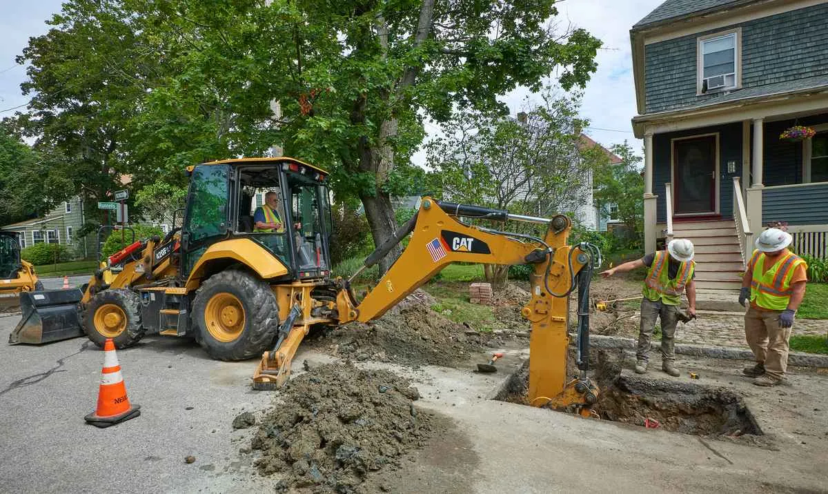 A yellow CAT backhoe loader is digging a utility trench in an asphalt road in a residential neighborhood. Two construction workers in hard hats and safety vests stand over the trench, while the backhoe operator sits inside the cab, all supervising the digging operation in front of a house.