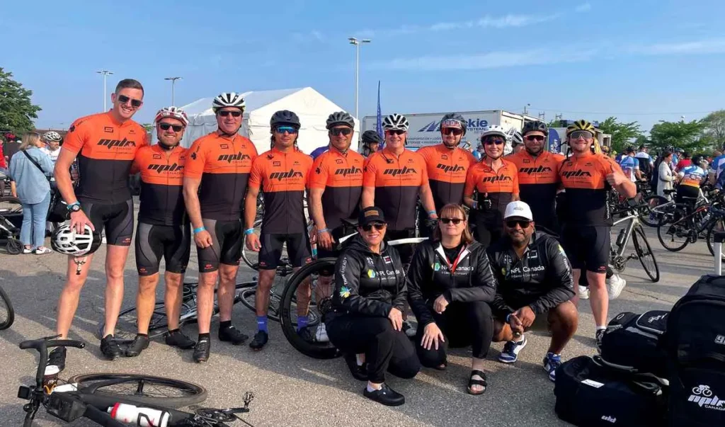A group of about twelve cyclists wearing matching orange and black cycling jerseys and helmets are posing for a photo at an outdoor cycling event. Three people in dark casual clothes are kneeling in the front.