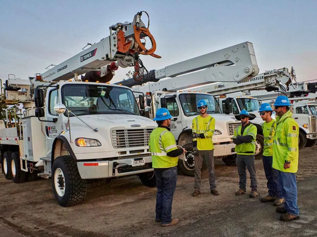 Five utility workers in hard hats and high-visibility vests are standing in a group meeting in front of a line of white utility and crane trucks, including a Terex Commander and an Altec vehicle.