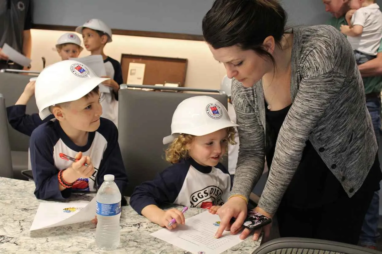 A woman is helping two young children wearing white hard hats and baseball shirts with a written activity or quiz at a table indoors, with other children visible in the background.