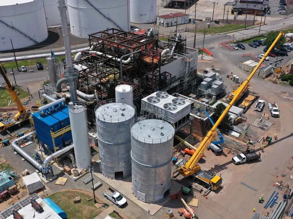 An aerial view of an industrial construction site with large silver storage silos, blue machinery, and a complex steel structure being built. A long yellow crane is visible near the construction area and white storage tanks are in the background.