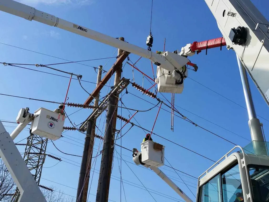Three utility workers in bucket trucks are performing maintenance high up on wood power poles and complex electrical wires against a clear blue sky. The truck booms are branded Altec.