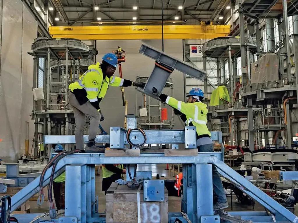 Two construction or industrial workers in blue hard hats and high-visibility vests are lifting and positioning a large steel component inside a spacious industrial facility with a yellow overhead crane.