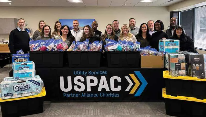 A group of approximately 16 Utility Services Partner Alliance Charities (USPAC) employees stands behind a table laden with donated items, including black tubs filled with hygiene kits and stacks of paper towels and napkins, inside a modern office space. A banner on the table reads 'Utility Services USPAC Partner Alliance Charities'.