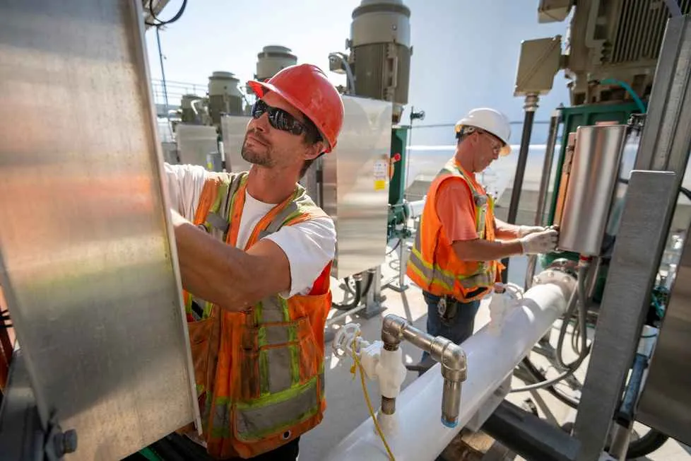 Two construction or plant workers in hard hats and high-visibility vests are performing maintenance on industrial machinery and pipes outdoors. The worker in the foreground wears a red hard hat and sunglasses.