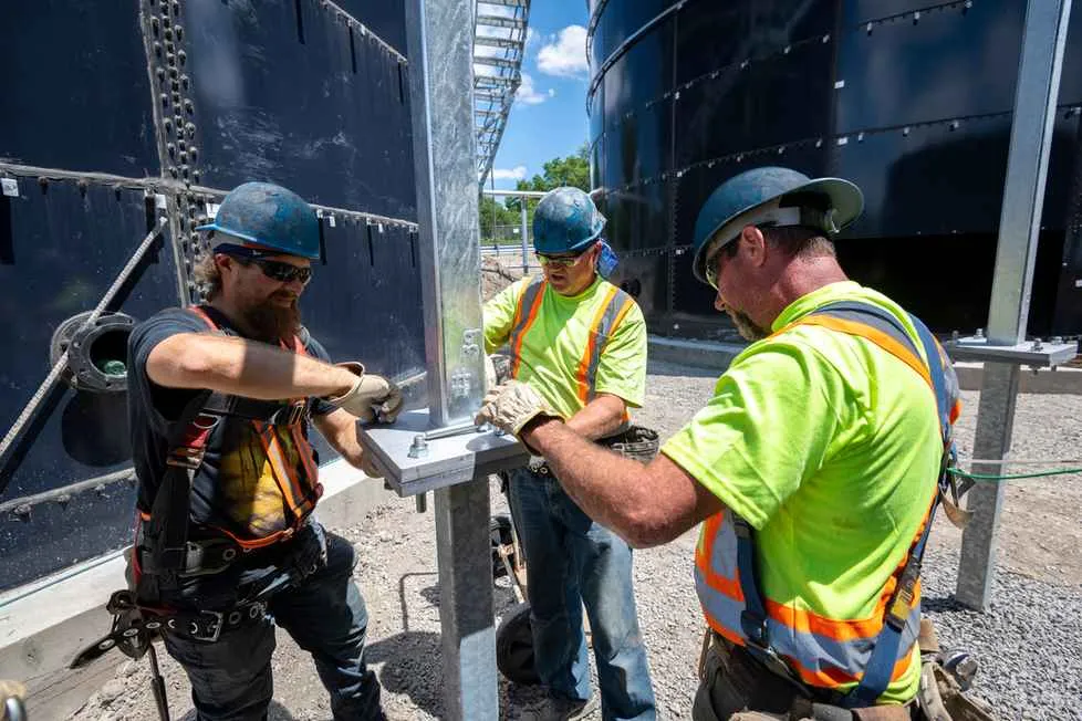 Three construction workers in hard hats and high-visibility gear are tightening bolts on a steel support beam near large, black industrial tanks.
