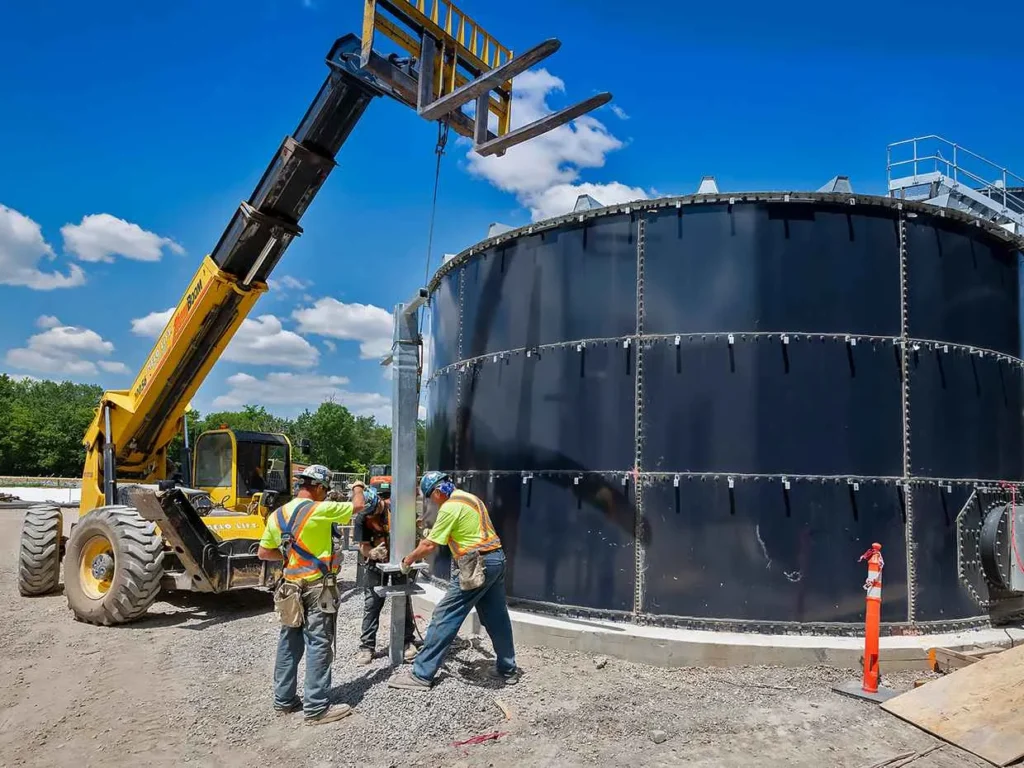 Construction workers are assembling a large, dark industrial storage tank outdoors, while a yellow telehandler or forklift is positioned nearby against a blue sky.