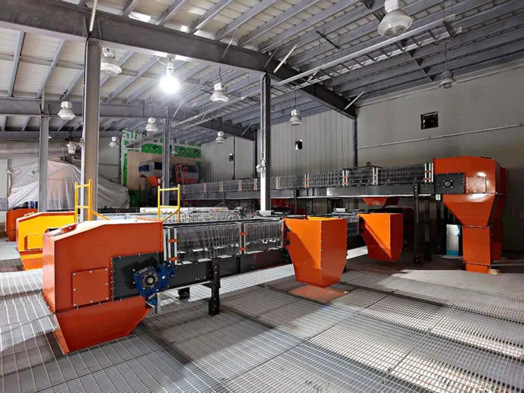 An interior view of a large industrial facility with a high ceiling, showing orange and black processing machinery sitting on a metal grate floor. The building structure is supported by steel beams.