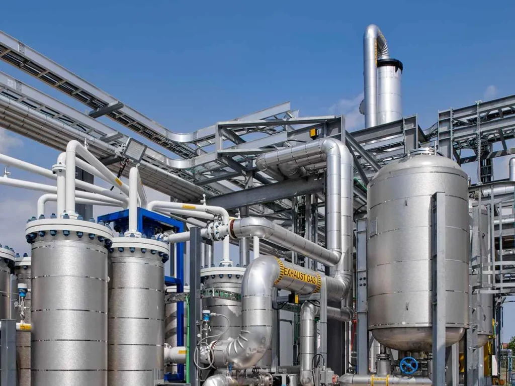 A large, complex industrial facility under a blue sky, featuring numerous shiny silver pipes, storage tanks, and structural steel frameworks. One large pipe is clearly labeled "EXHAUST GAS".
