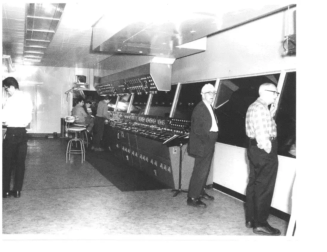 A black-and-white, vertically oriented photo showing men standing in an industrial control room with a long console full of switches and buttons.