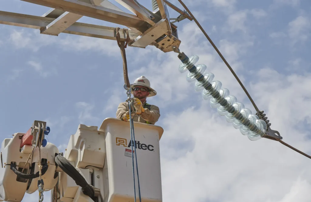 A utility lineman wearing a hard hat and safety glasses works from a bucket truck on an electrical power line.