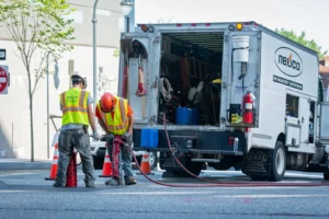 Two construction workers are on a street. One worker, wearing a hard hat and a high-visibility vest, is operating a jackhammer on the asphalt. Another worker, also in a hard hat and high-visibility vest, stands nearby.