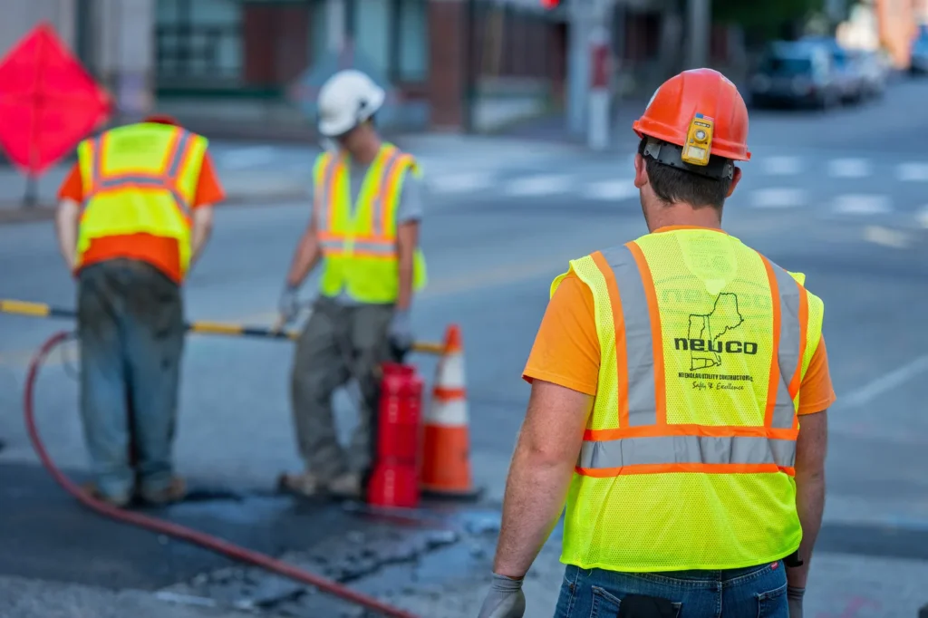 A construction worker wearing an orange hard hat, orange shirt, and a yellow high-visibility vest with the "neuco" logo on the back is shown from behind, looking at two other workers in the background.