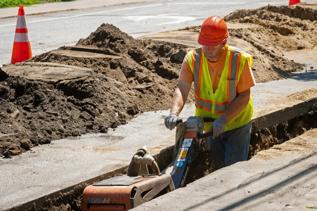 A man in a hard hat, safety vest, and sunglasses is kneeling on a city sidewalk, working on a piece of equipment attached to large black pipes.