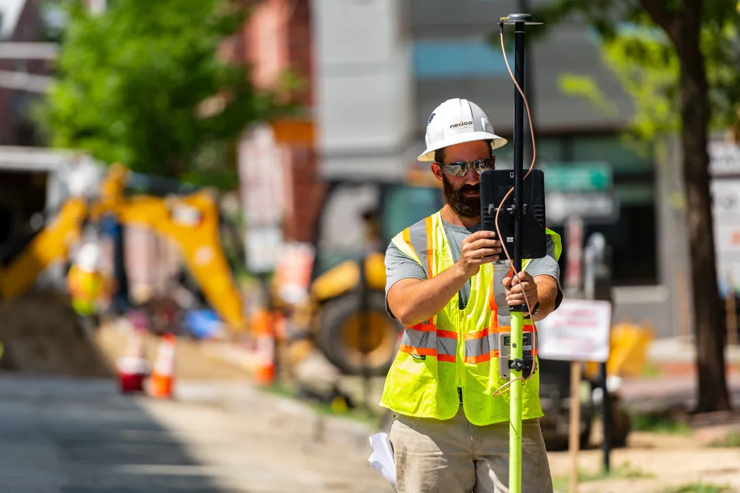 A man wearing a hard hat and a high-visibility vest is holding a piece of surveying equipment on a construction site.
