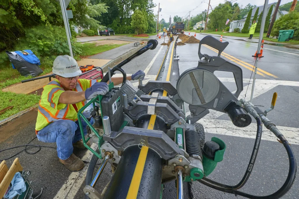 A construction worker wearing a hard hat and a high-visibility vest is kneeling on a street, operating a fusion welding machine to join a large yellow-striped plastic pipe.