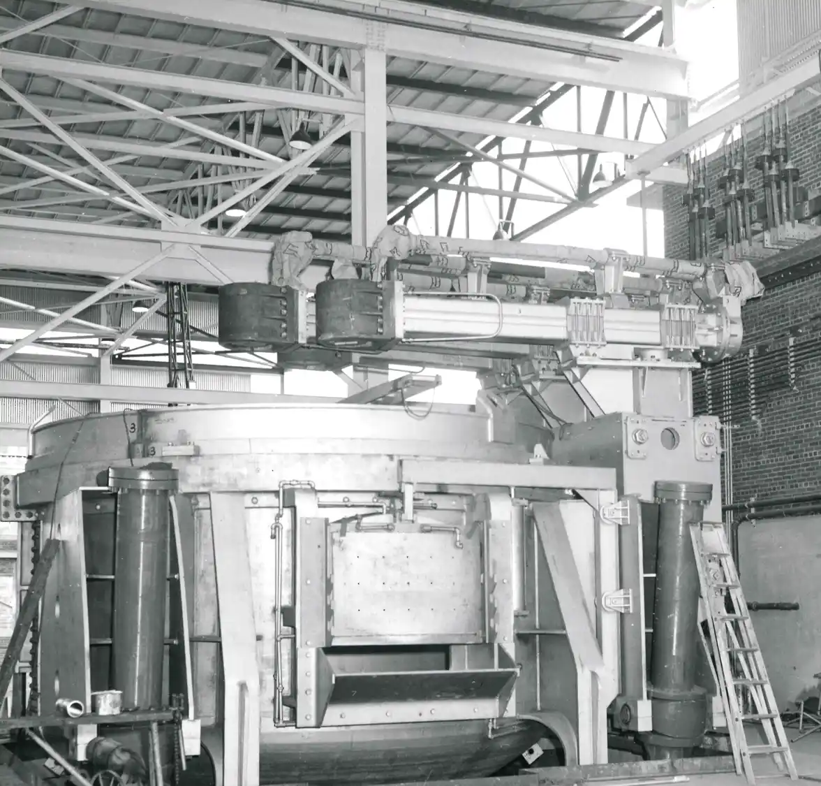 A black-and-white, interior photograph of a large, heavy industrial machine or furnace with complex piping and overhead structural steel beams, likely taken in Perth Amboy in 1957.