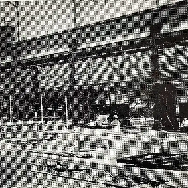 A grainy, black-and-white photo shows a construction or manufacturing scene inside a large building, with a prominent steel I-beam structure supported by columns, and workers moving equipment on the floor below.