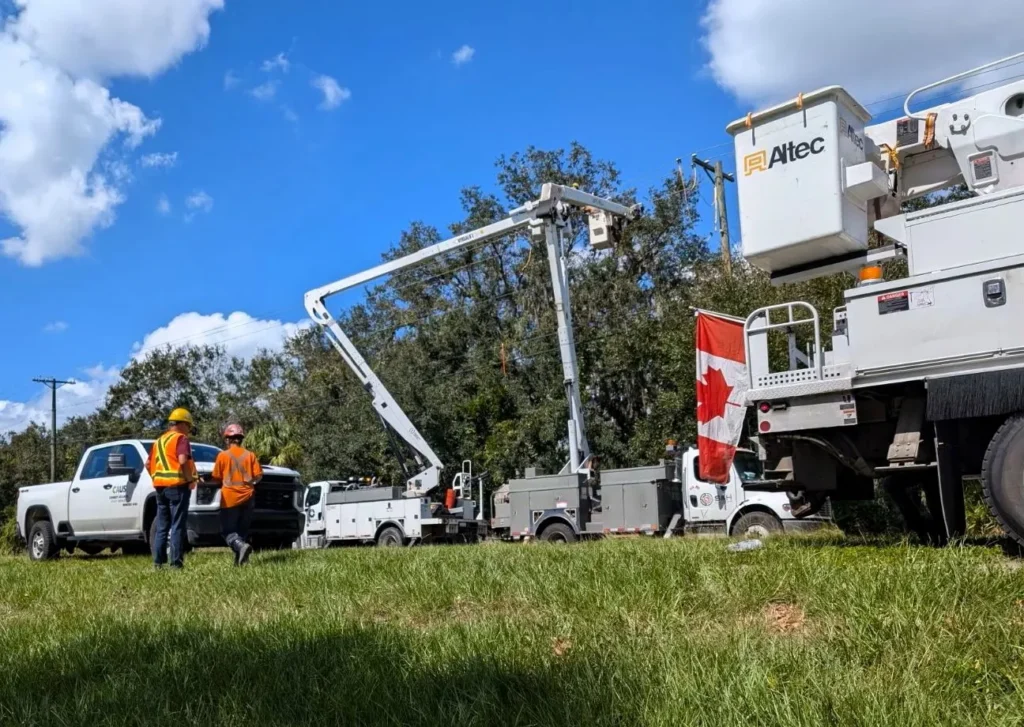 Two utility workers in safety gear stand next to several Altec bucket trucks and equipment performing power line work on a sunny day, with a Canadian flag visible on one vehicle.