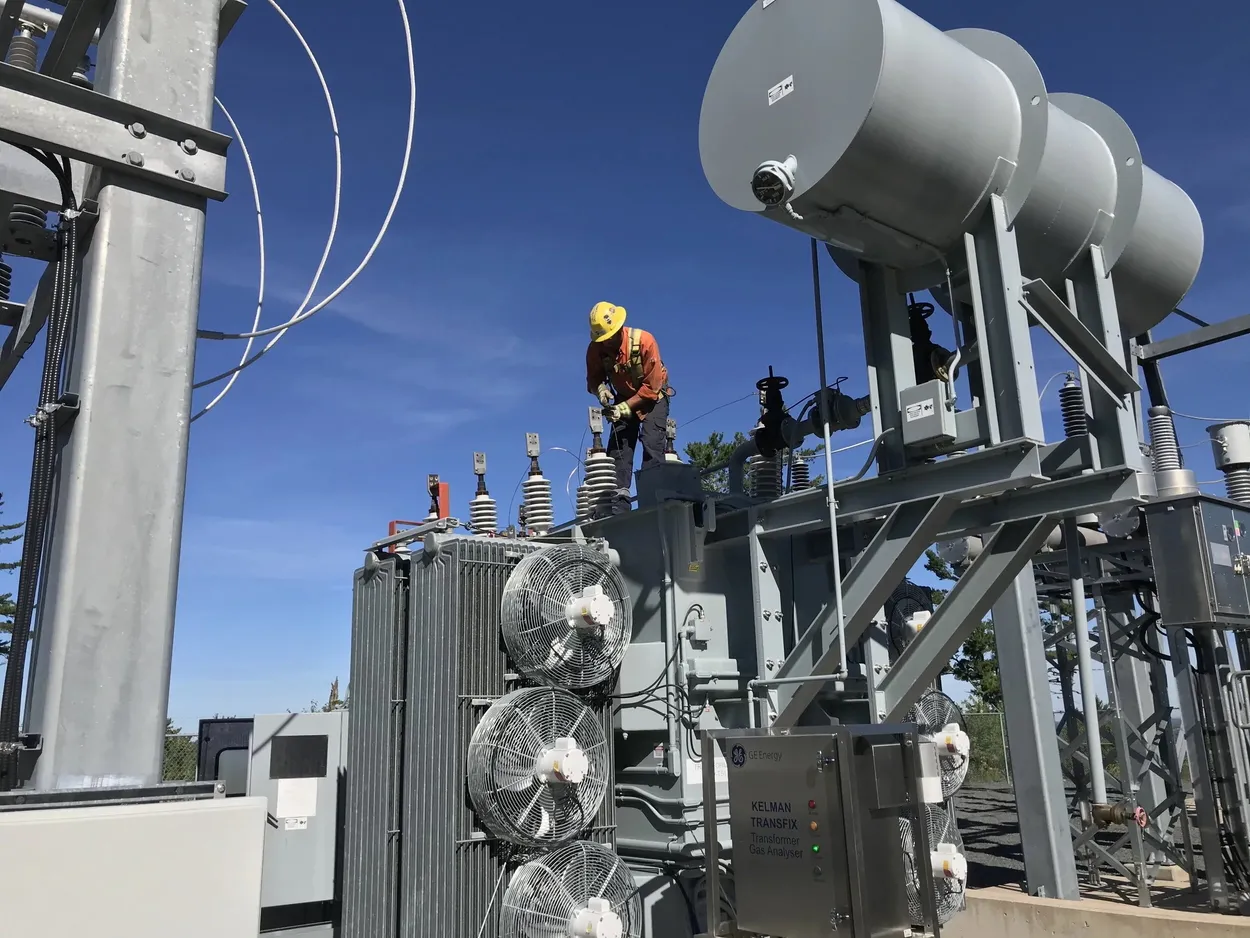 An electrical worker in safety gear performs maintenance on a large power transformer at an outdoor substation.