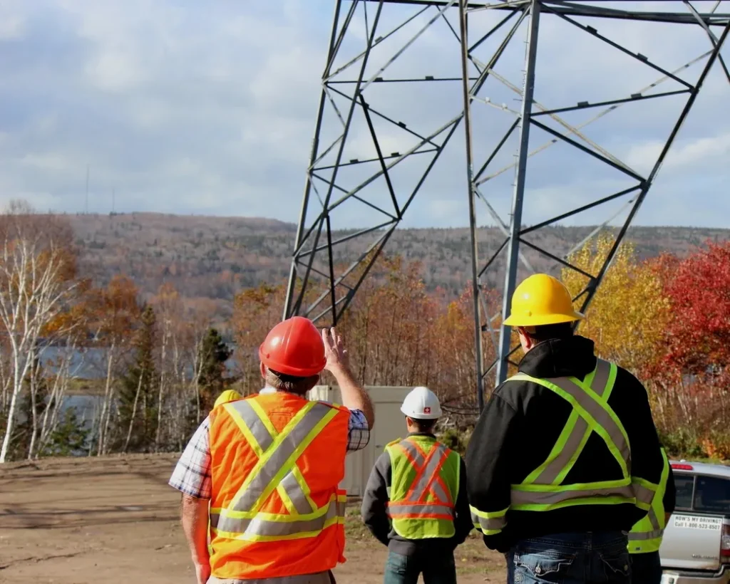 Three construction workers in safety vests and hard hats stand near a large electrical transmission tower, looking at a forested landscape.