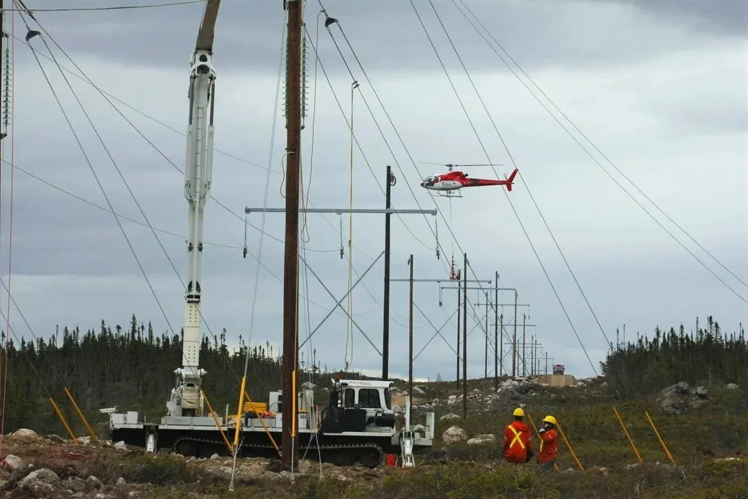Construction workers and a helicopter near new power transmission poles in a rugged landscape.