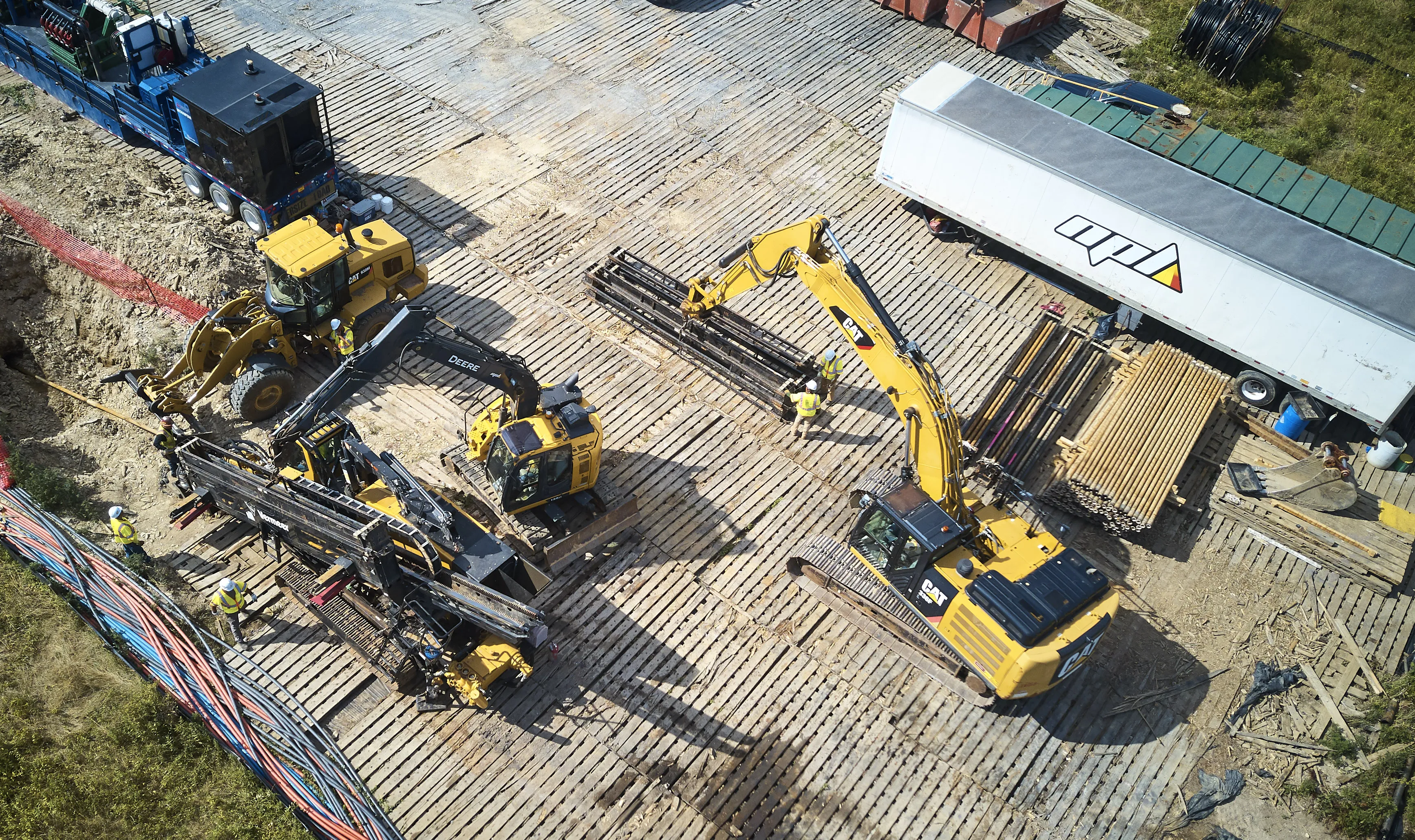 An aerial view of a construction site shows several pieces of heavy machinery, including a yellow CAT excavator and a wheel loader, operating on a platform of wooden mats.